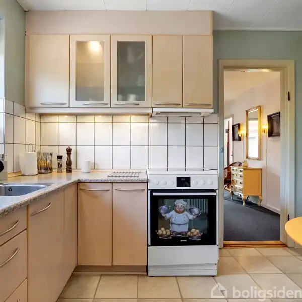 Et lyst køkken med beige skabe og fliser. En ovn står under en kogeplade, omgivet af en vask og arbejdsflader. Et åbent dørhul fører til en gang.