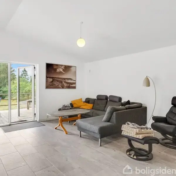 En stue med en grå sofa og lænestol oplyst af en gulvlampe. Glasdøre fører ud til en terrasse med udsigt til træer. Billedet viser også et maleri på væggen.