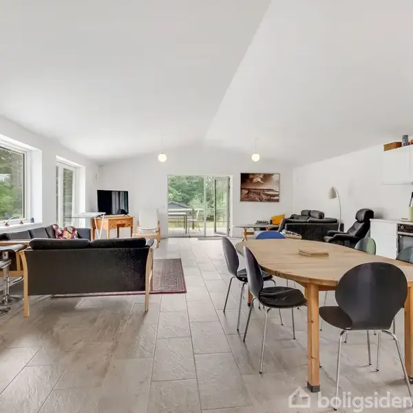 En rummelig stue-køkken alrum med lyst trægulv, sorte og grønne stole om et spisebord, sorte sofaer nær vindue, og åbne hylder i køkkenet. Store vinduer giver udsigt til natur.