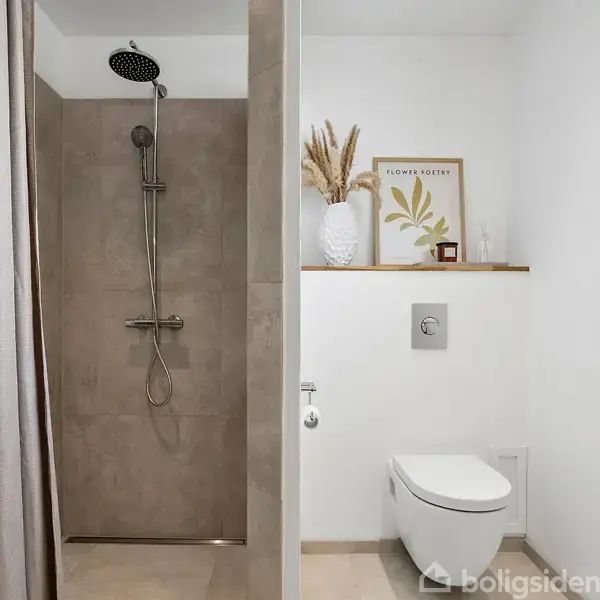 Bruseområde med grå fliser og en rund bruser. Til højre hænger et toilet på væggen, og over det står en vase og et indrammet billede med teksten "FLOWER POSTER."