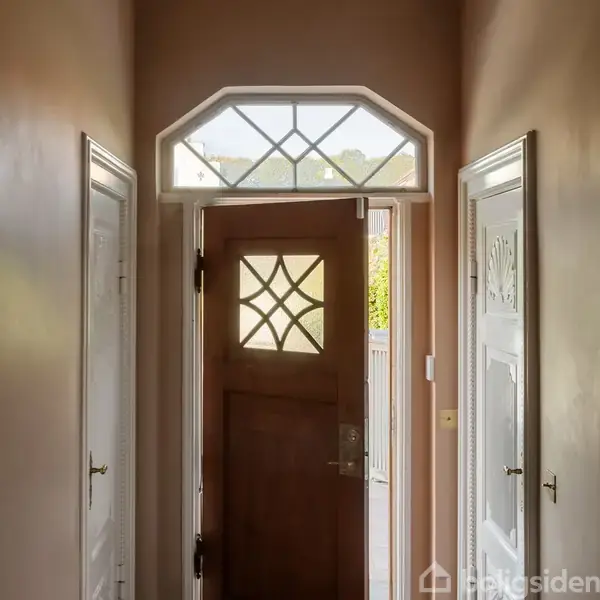 Trædør med geometriske glasindlæg står åben, og lukker lys ind i en gang med lyserøde vægge og hvide dørkarme. Indgangen udviser klassisk design med symmetriske detaljer.