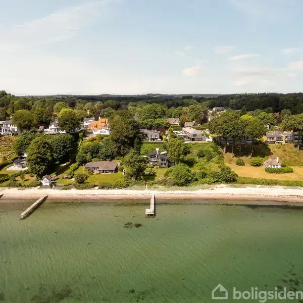 Kystnære huse står stille på en frodig, grøn bakke med udsigt over en klar vandfyldt bugt. Træer omkranser området og små badebroer strækker sig ud i vandet.