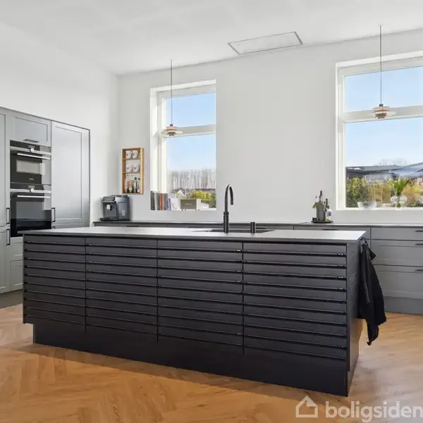 Køkken med mørke skabe og ø midt i et lyst rum. To vinduer giver udsigt og lys, indrammet af hvide vægge. Trægulv og moderne apparater supplerer indretningen.