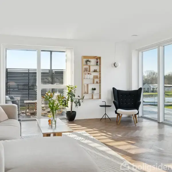 En lys stue med sofa og lænestol placeret ved store vinduer. Et sofabord står centralt med planter ovenpå. Væggene er hvide, og der er udsigt til udendørsområder.