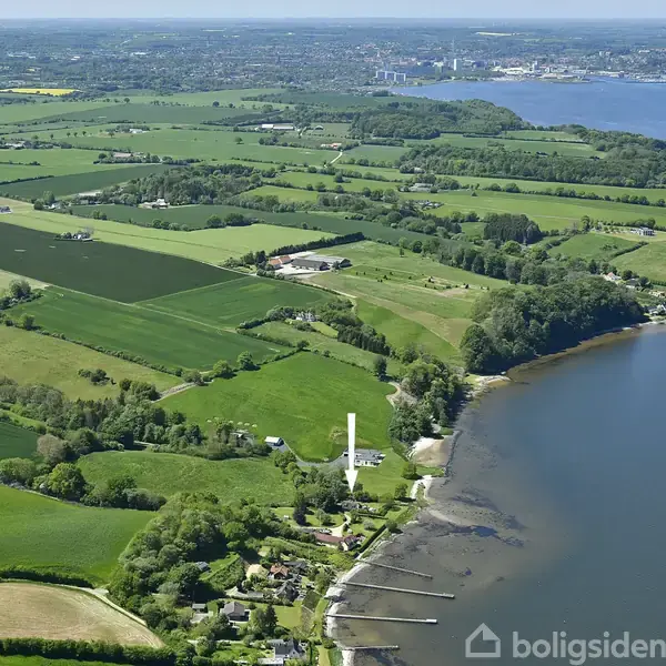 Landskab set fra luften med grønne marker, en hvid mølle i centrum, omgivet af huse. I baggrunden ses en bugt med vand og en fjerntliggende by.