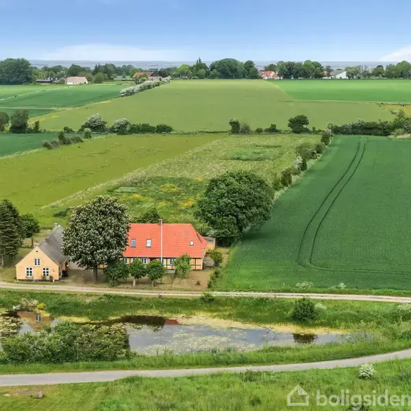 Gule bygninger med rødt tag ligger ved en lille vej. Omgivet af grønne marker og træer i et landligt område.