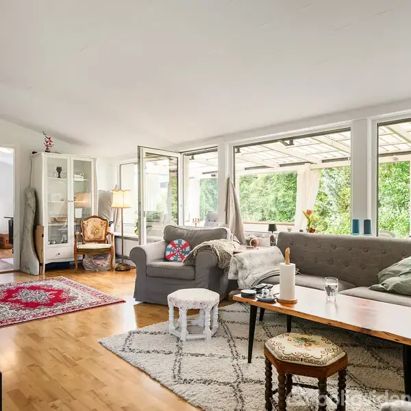 Stue med grå sofa og sofabord, dekoreret med puder og et tæppe. Rummet er lyst med store vinduer, der åbner ud mod en terrasse med grøn udsigt.