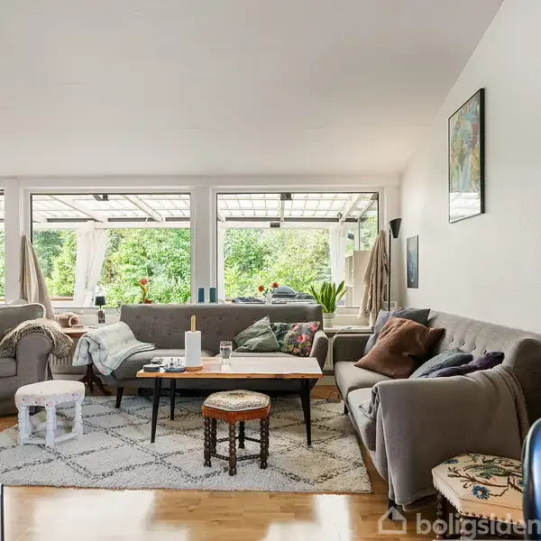 Stue med to sofaer omkring et sofabord; rummet har store vinduer, der giver udsigt til en grøn have. Der er tæppe, puder og maleri på væggen.