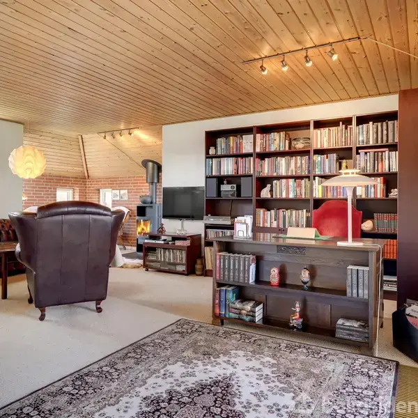 Et bibliotek med en stor bogreol fyldt med bøger, lænestole i midten, og en tæppebelagt gulv. Rummet har træloft, store vinduer og moderne belysning.