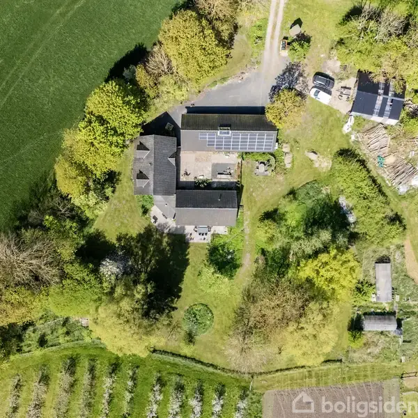 Hus med solpaneler ligger i frodig, landlig omgivelser omgivet af træer og marker. Flere små bygninger og parkerede biler ses nær hovedbygningen. En sti fører væk fra ejendommen.