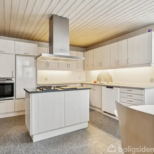 Et moderne køkken har hvidt interiør med en central kogeø og indbygningsovn. Omgivet af overskabe, et vindue til højre, grå gulvfliser og en rund spisebord med stole.