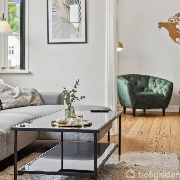 Et udendørs gårdmiljø med et bord og stole er placeret på en flisebelagt terrasse, omgivet af en hvid mur og to bygninger med murstensfacade og rødt tegltag.