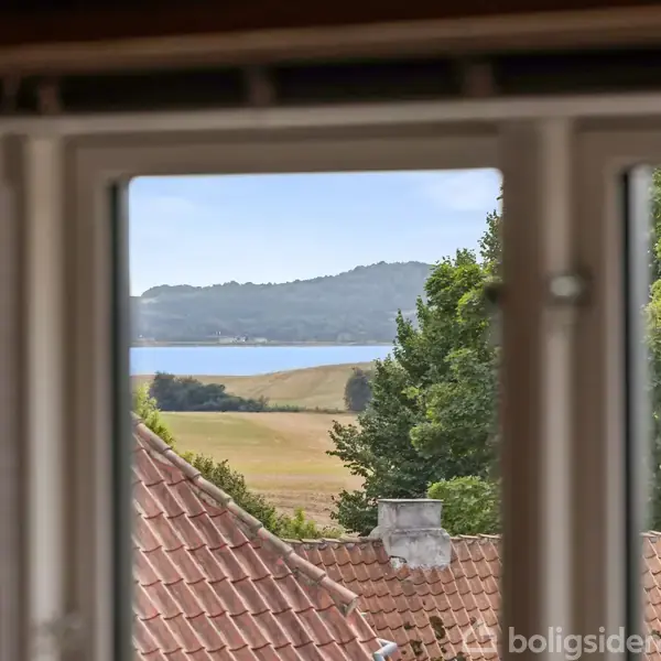 Et vindue i forgrunden viser et landskab med røde tagstenstage, træer og en sø eller fjord i baggrunden, omgivet af bakket terræn og skov.