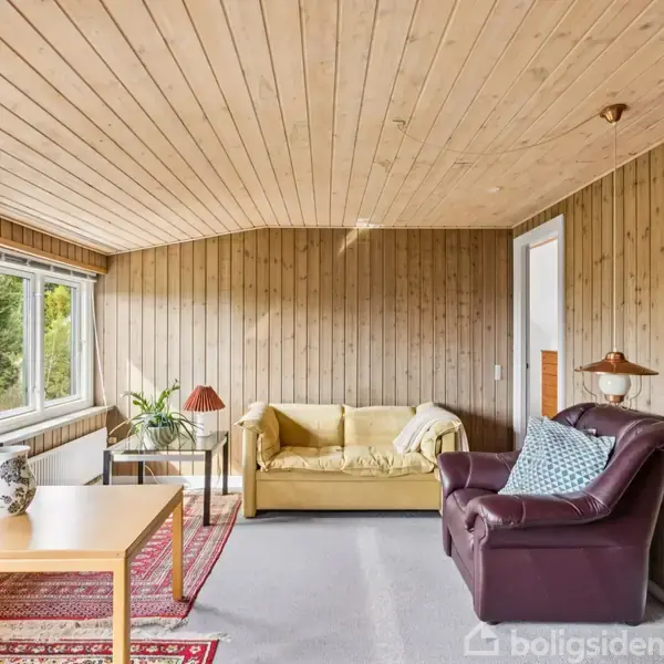 En stue med en gul sofa og en brun lænestol arrangeret på en grå tæppebelagt gulv. Væggene og loftet er af træpaneler. Et stort vindue slipper lys ind.