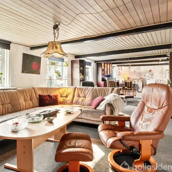 Brun lædersofa og matchende lænestol står i solbelyst stue, omgivet af træloft og tæppebelagt gulv. Kaffe- og spisebord i baggrunden skaber en hyggelig atmosfære.