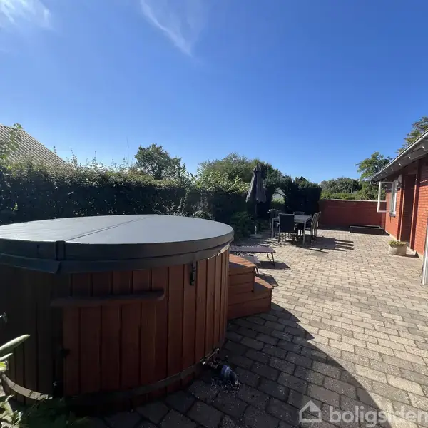 En rund, overdækket udendørs jacuzzi står på en flisebelagt terrasse. Omkring den er havemøbler og en rød murstensbygning under en blå himmel omgivet af grønne buske og træer.