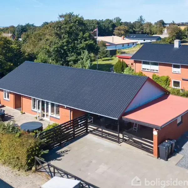 En rød murstensvilla med sort tag står stille omgivet af en flisebelagt terrasse og grønne buske. Baggrunden viser flere huse og frodige træer under en klar blå himmel.