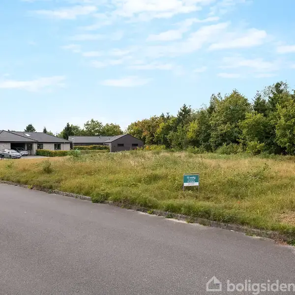 Grunden ligger ubenyttet, beliggende ved asfalteret vej omgivet af huse og træer. Et skilt på grunden viser teksten "TIL SALG".