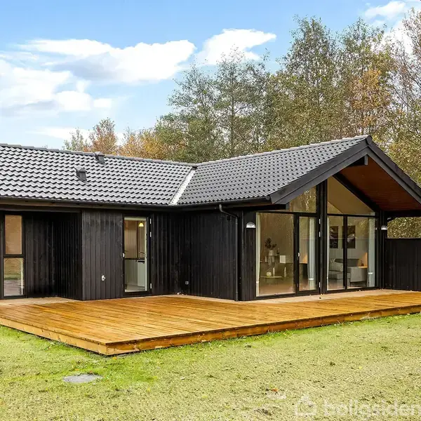 Sort træhus står stille på græsklædt grund med stor træterrasse foran store vinduer. Omkring huset er der træer og blå himmel i baggrunden.