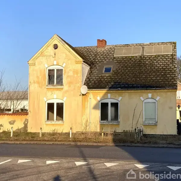 Gul, ældre to-etagers bygning med skrånende tag og solpaneler, står ved en vej på en solrig dag. Vegetation omkring bygningen.