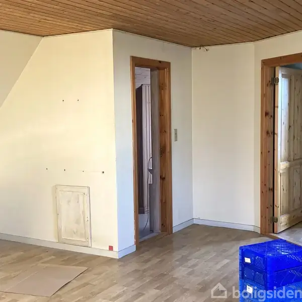 Et tomt, lyst rum med trægulv og bræddevægge; en blå plastkasse står på gulvet med adgang til to åbne døråbninger, der fører til tilstødende rum.