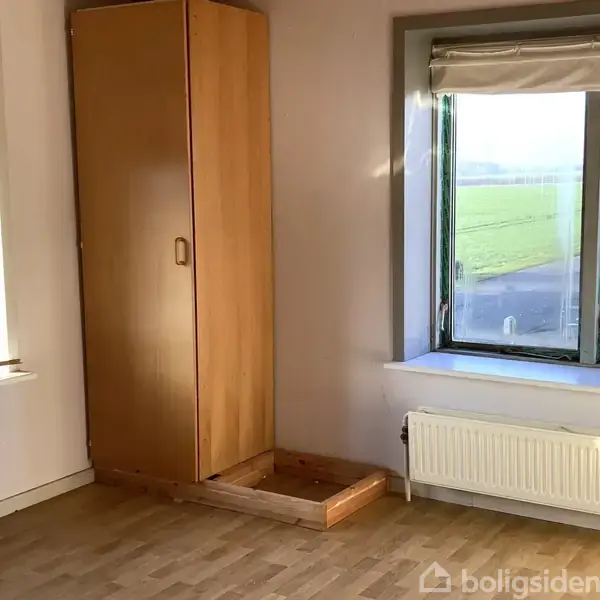 Et træskab står åben foran et stort vindue med udsigt til en mark. Rummet har lyse vægge, en radiator under vinduet og trægulv.