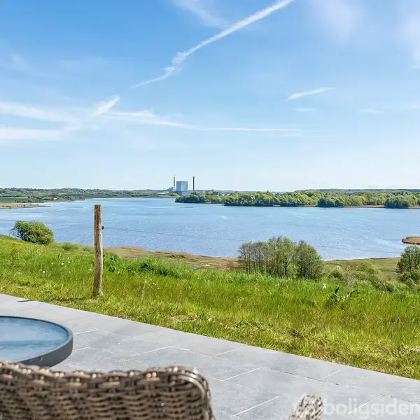 Et rundt bord med planter på står på en terrasse, der har udsigt over en sø omgivet af grønne enge og skov under en klar blå himmel.