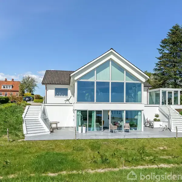 Hus med store glasfacader og trekantet gavl på en åben, grøn græsplæne. Trapper på begge sider fører til terrassen. Baggrund med blå himmel, træer og nabo hus.