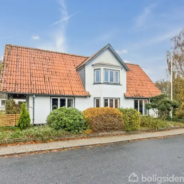 Hvidt hus med rødt tegltag og en kvist, omgivet af buske og træer, står ved en asfalteret vej. Huset har flere vinduer og en indkørsel til venstre.