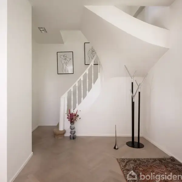 Trappeopgang med hvidmalet trappe fører opad, omkranset af hvide vægge med to kunstneriske billeder. En vase med blomster står ved trappen, og en frakkestander står ved siden af. En vindue åbner op for naturligt lys.