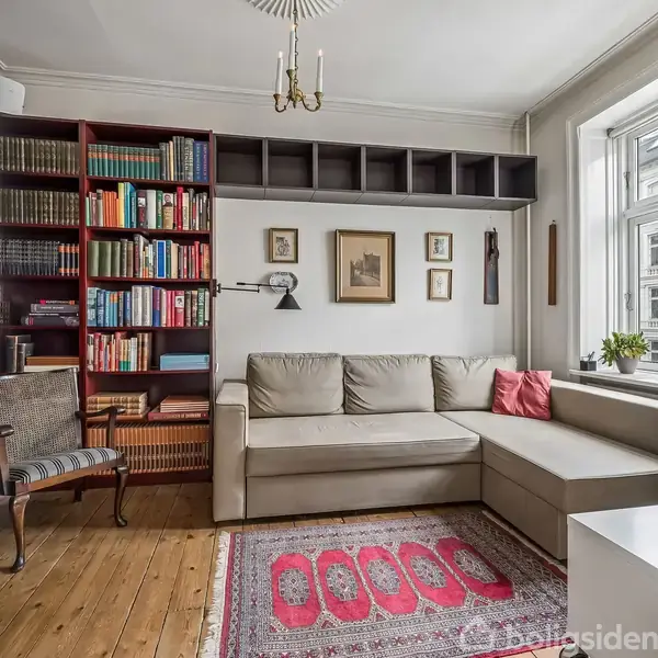 En stue med grå hjørnesofa, flankeret af bogreoler fyldt med bøger. En rød pude og tæppe tilfører farve. Et vindue til højre giver lys, omgivet af rammede billeder.