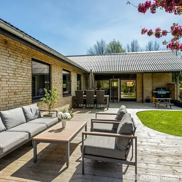 Grå sofaer og bord står på en træterrasse. I baggrunden ses et spisebord og græsplæne med blomstrende træ, under en klar blå himmel.