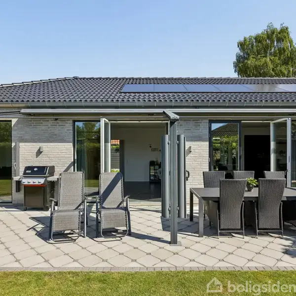 En moderne terrasse med spisebord og stole står i sollys. En grill er placeret ved husets åbne glasdøre, omkranset af en grøn have og et lysgråt hus.