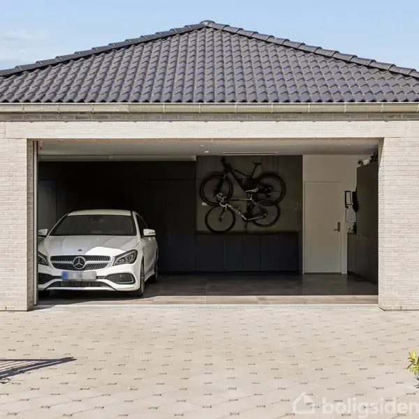 Hvid bil parkeret i åben garage med fladt, mørkt tag. To cykler hænger på væggen. Der står "20" på højre side af bygningen.
