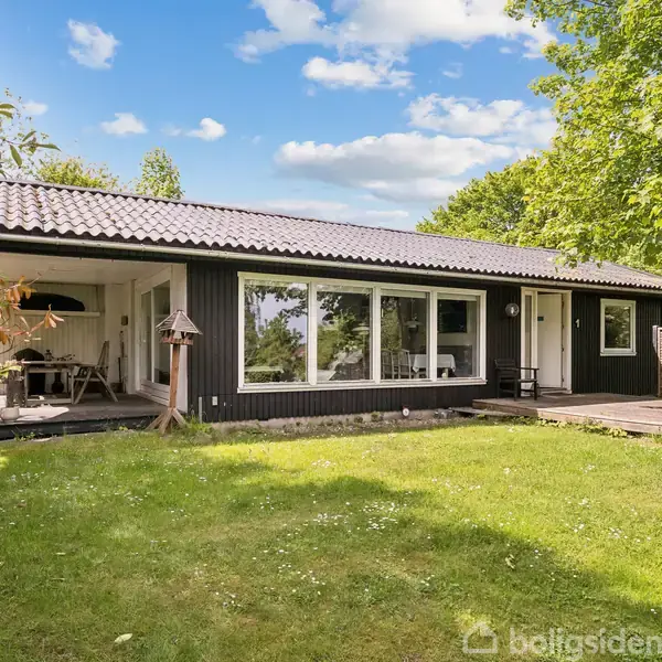 Et sortmalet enfamiliehus med store vinduer står i en grøn have. En overdækket terrasse til venstre indeholder havemøbler. Solen skinner, og træer omkranser området.
