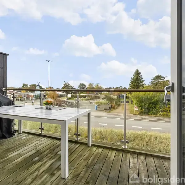 En hvid bord står på en træterrasse med glasrækværk, ved siden af en grill med betræk. En vej og grønne træer ses i baggrunden under en blå himmel.