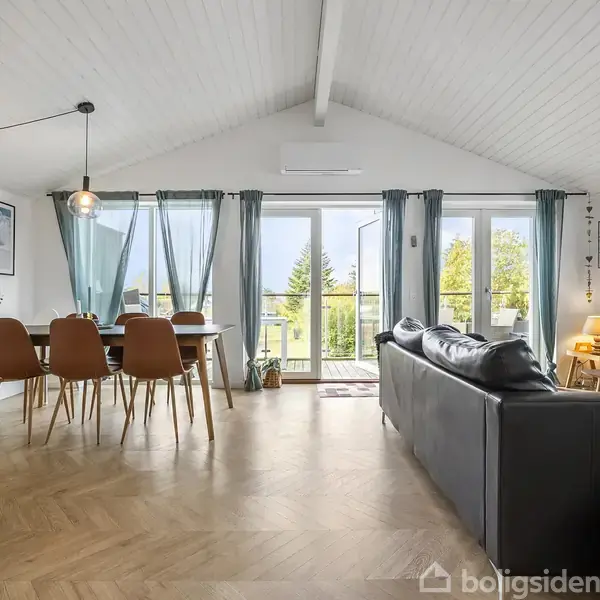 Stue med spisebord og stole til venstre, sofa til højre. Lyseblå gardiner hænger foran store vinduer med udsigt til have. Trægulv og hvide vægge.