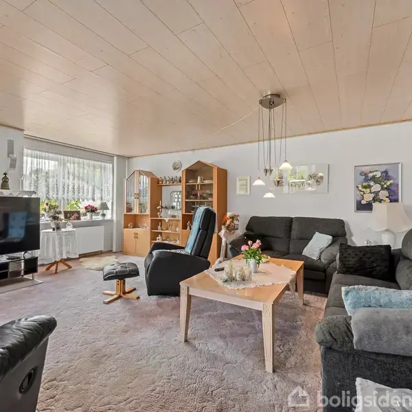 Et rummeligt, tæppebelagt stueområde med en stor, mørk sofagruppe og lænestol omkring et træbord, dekoreret med blomster. En stor fladskærms-tv står ved et vindue med et skab i hjørnet.