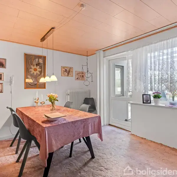 Spisebord dækket med lyserødt dug; omgivet af sorte stole. På bordet står en vase med blomster. Rummet har billeder på væggen og store vinduer med gardiner, der lukker lys ind.