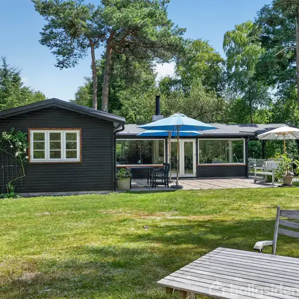 Et sortmalet sommerhus med store vinduer står i en grøn have, omgivet af træer. Et havebord og stole er placeret på græsplænen under en blå parasol ved siden af terrassen.