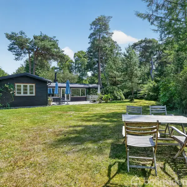 Et mindre sommerhus står i en grøn have omgivet af træer. Der er et træbord med fire stole i forgrunden. På terrassen ses parasoller.