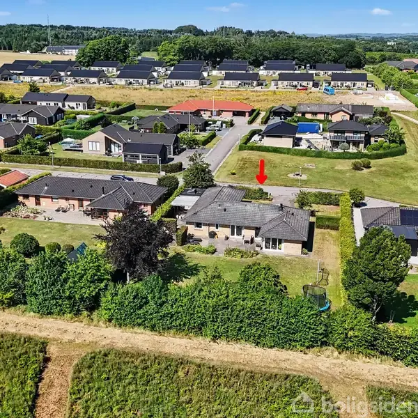 Hus markeres med rød pil, beliggende i et boligområde med flere huse og grønne haver, omkranset af natur og marker i baggrunden.