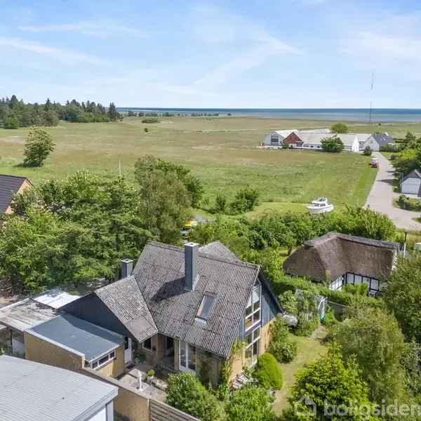 Et hus med skråtag står omgivet af træer og andre mindre huse. Baggrunden viser en stor græsmark, en vej og fjerntliggende bygninger nær kysten.