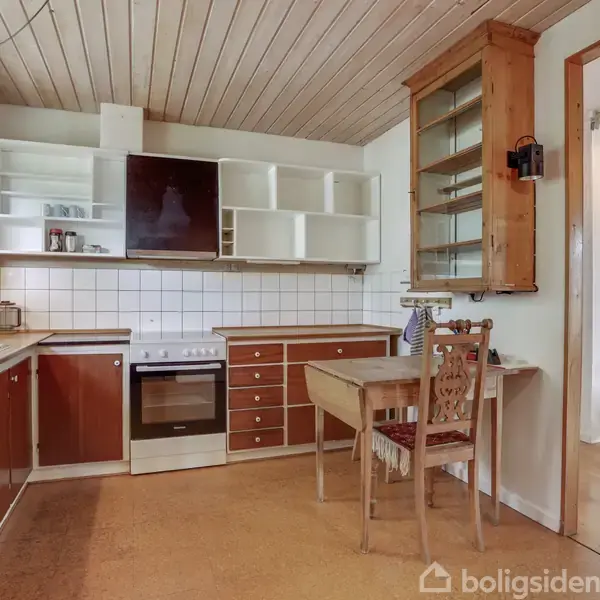 Et køkken med brune skabe har en ovn i midten. En træstol står ved et lille bord. Omkring er der fliser på væggen og åbne hylder.