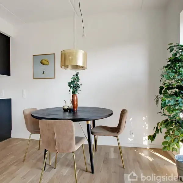Rundt spisebord med tre stole i en lys spisestue. En gulvplante står ved siden af det store vindue, og et maleri hænger på væggen. En gylden lampe hænger over bordet.