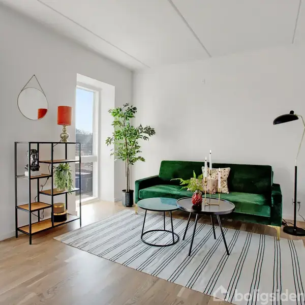 Stue med grøn sofa og sofabord på stribet tæppe, omgivet af gulvlampe, reol med planter og to store vinduer, der lader naturligt lys strømme ind.