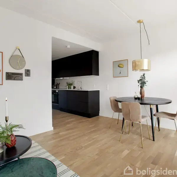 Et moderne spiserum med et rundt bord og fire stole står i forgrunden. En lys pendellampe hænger over bordet. Køkkenet ses i baggrunden med mørke skabe.