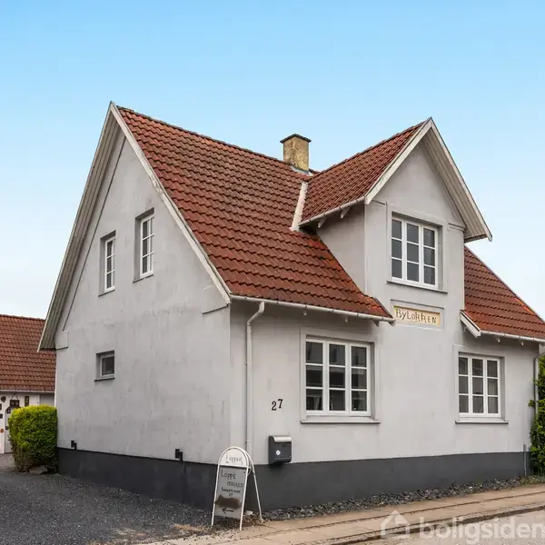 Huset er en hvid muret bolig med rødt tegltag. Det står stille på en gade med have og en indkørsel. Nummeret 27 er synligt, omgivet af andre huse.