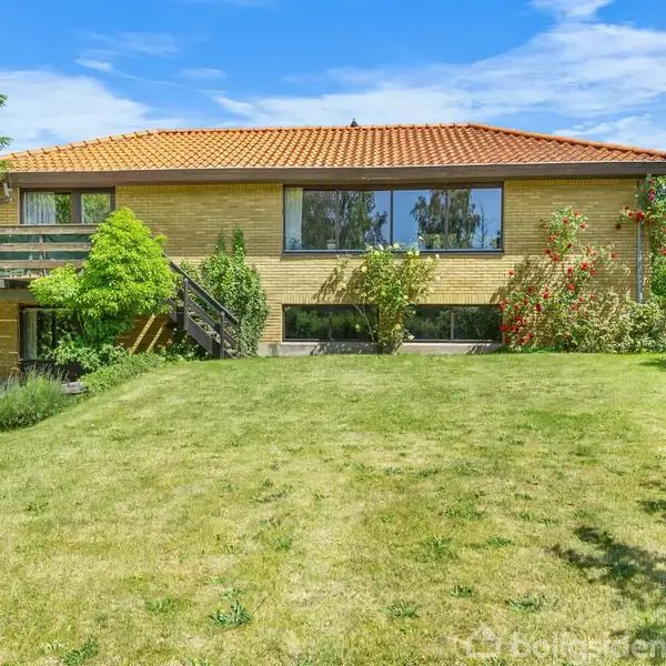 Et hus med rødt tegltag står på en velholdt græsplæne omgivet af buske og træer. Sollys oplyser facaden, og en balkon ses til venstre.