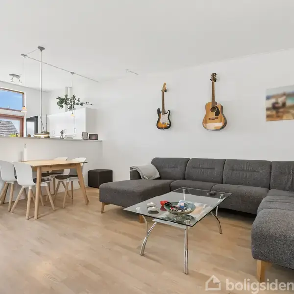 En moderne stue med en grå, L-formet sofa. Et glasbord står foran sofaen. To guitarer hænger på væggen. Et spisebord med stole er i baggrunden.